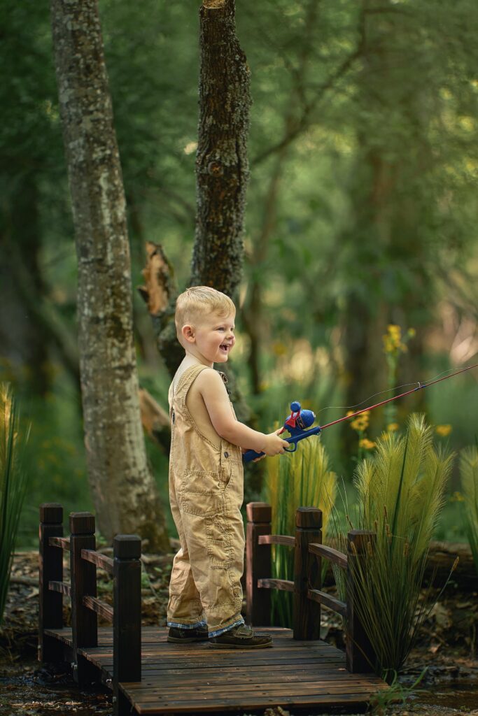 toddler three year old boy in khaki overalls holding fishing rod on bridge and laughing